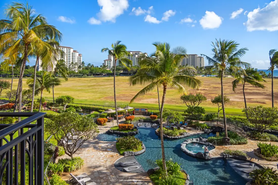 Una zona de piscina al aire libre ajardinada, con palmeras y edificios al fondo.