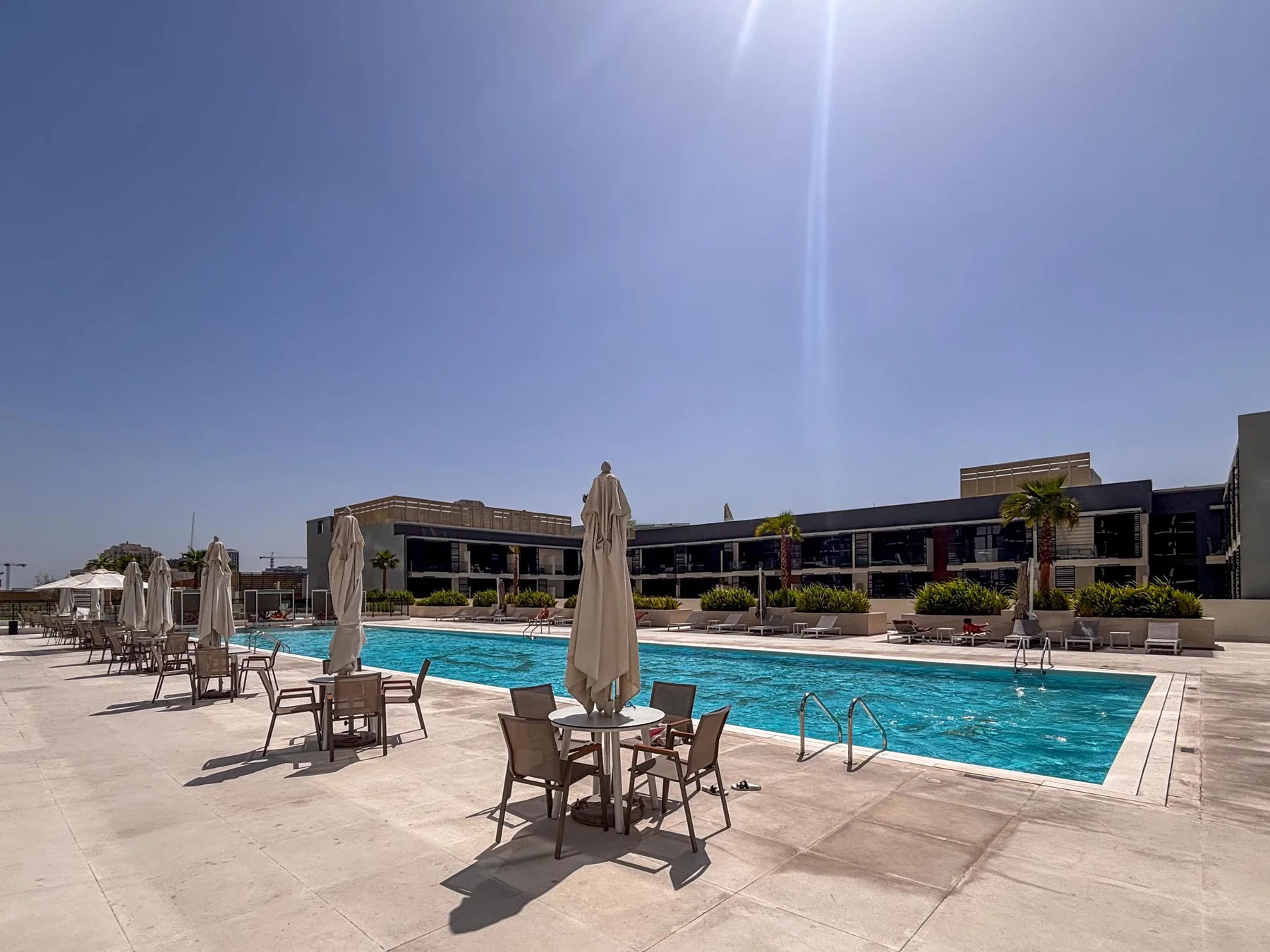 An outdoor swimming pool beside a modern low-rise building, with tables and seating on the deck.