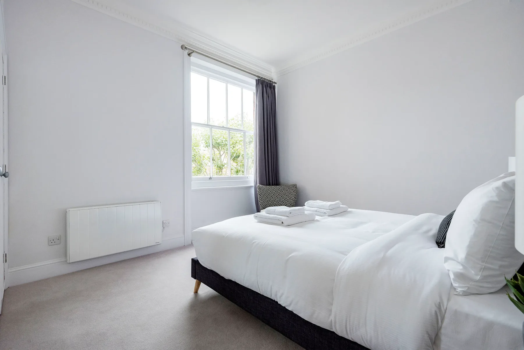 A bright bedroom with white bedding and a window overlooking greenery.