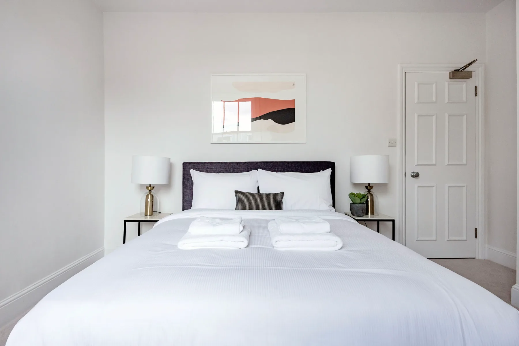 A neatly made bedroom with white bedding, bedside lamps, and framed artwork above the bed.