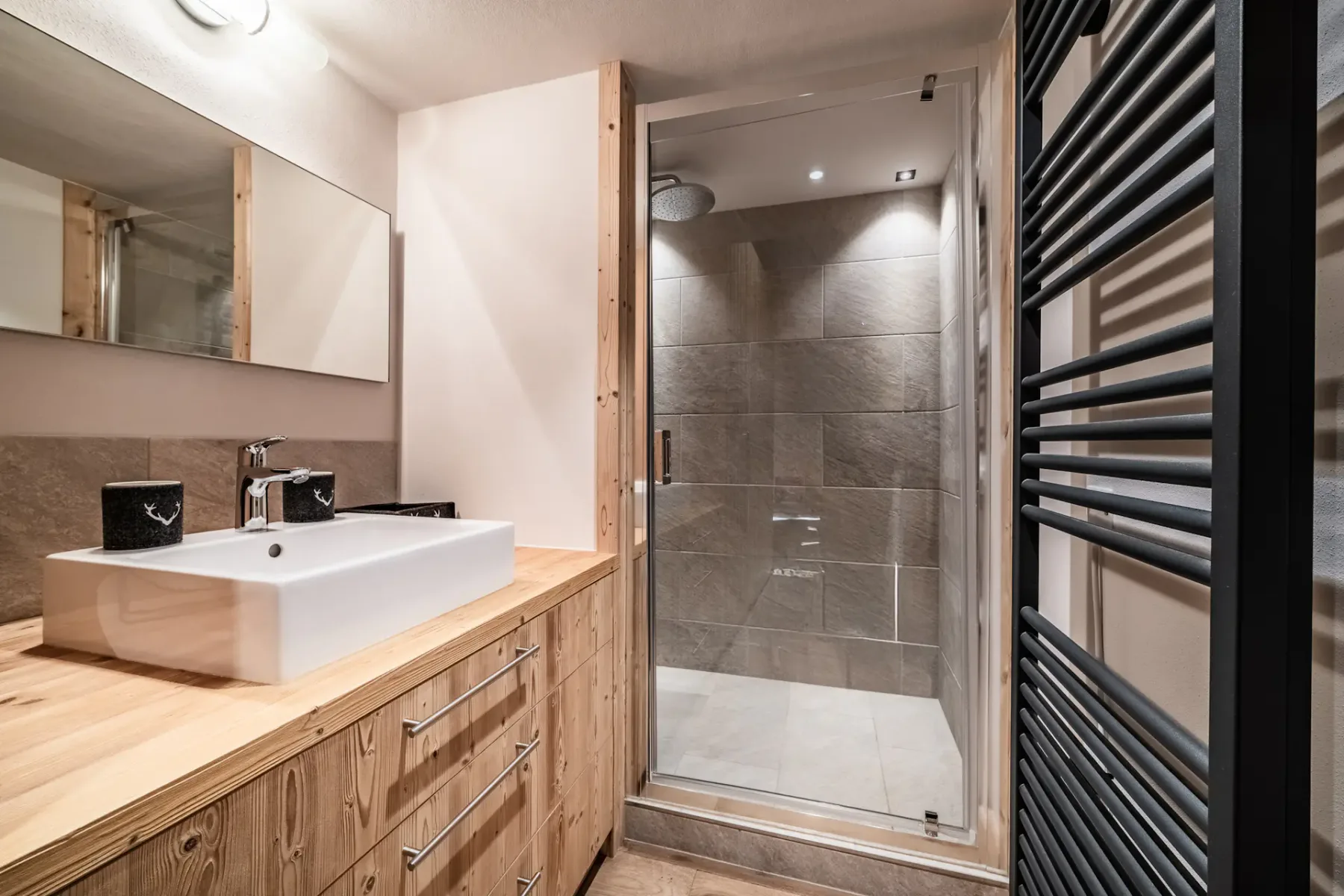 Modern bathroom with a wooden vanity, white rectangular sink, large mirror, glass-enclosed shower, and black towel radiator.