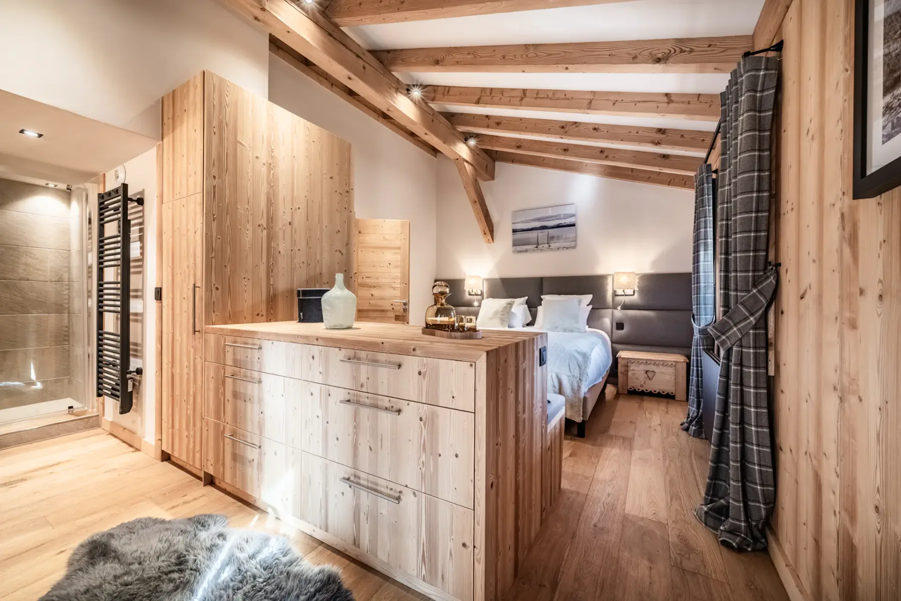 Cozy bedroom with wooden walls, a double bed with white bedding, a painting above the headboard, and a window with plaid curtains.