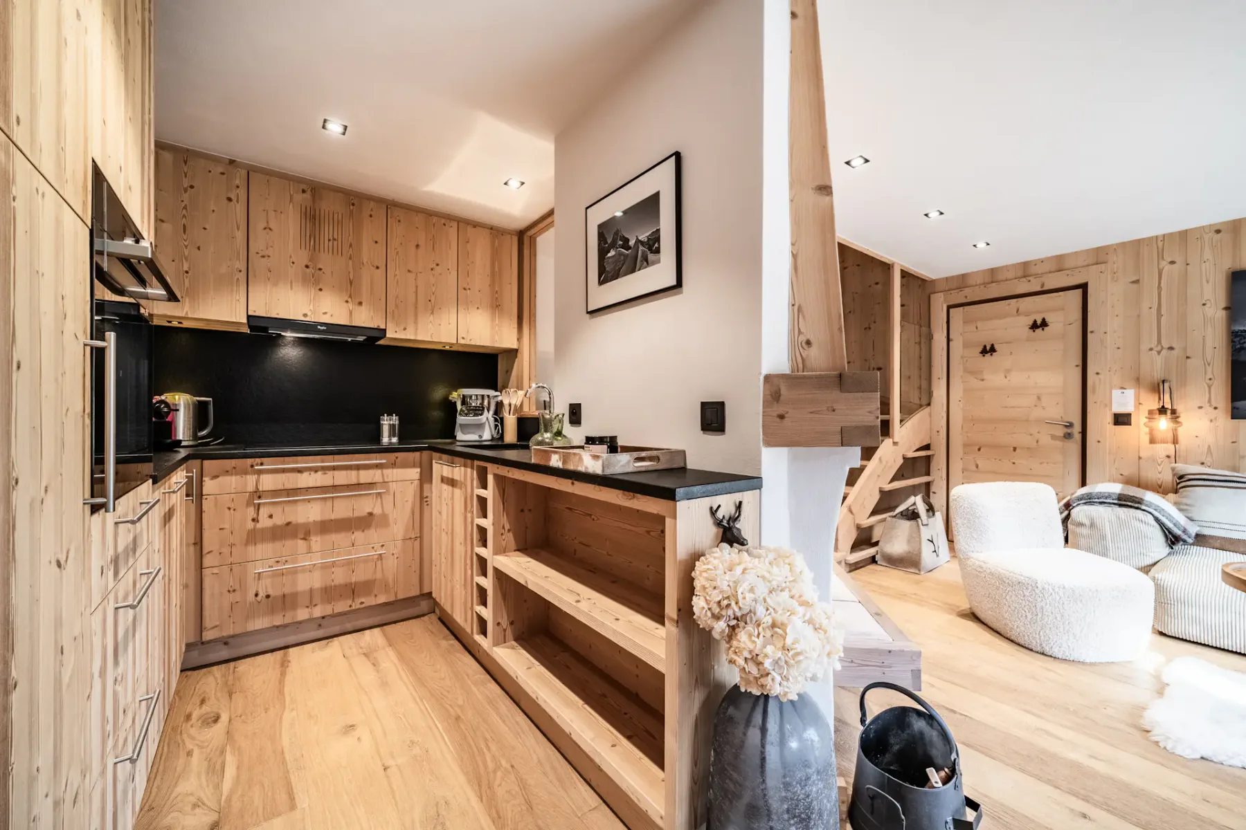 Modern kitchen with wooden cabinets, black countertops, and cozy seating area. Soft lighting and rustic decor create a warm, inviting atmosphere.