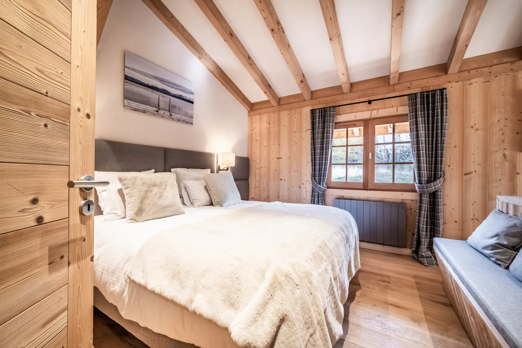 Cozy bedroom with wooden walls, a double bed with white bedding, a painting above the headboard, and a window with plaid curtains.