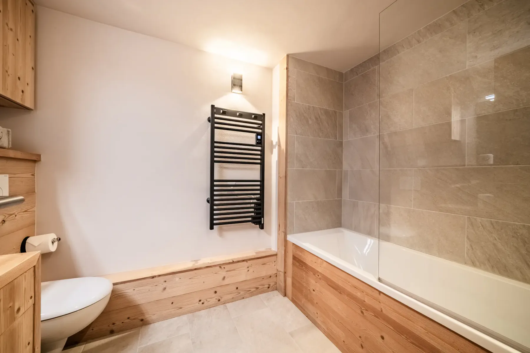 Modern bathroom with wooden accents, featuring a bathtub with glass screen, wall-mounted towel rack, and toilet on a tiled floor.