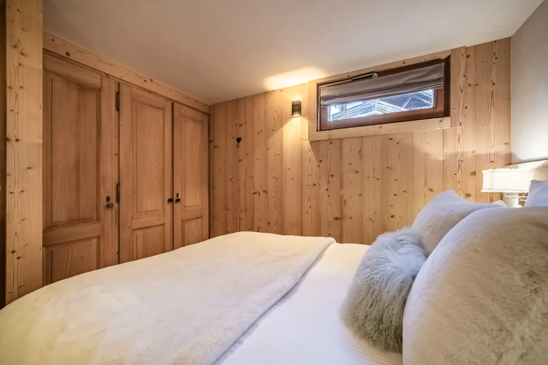 Cozy bedroom with a double bed, wooden walls, soft lighting, and a small window. A nightstand with a lamp is beside the bed.
