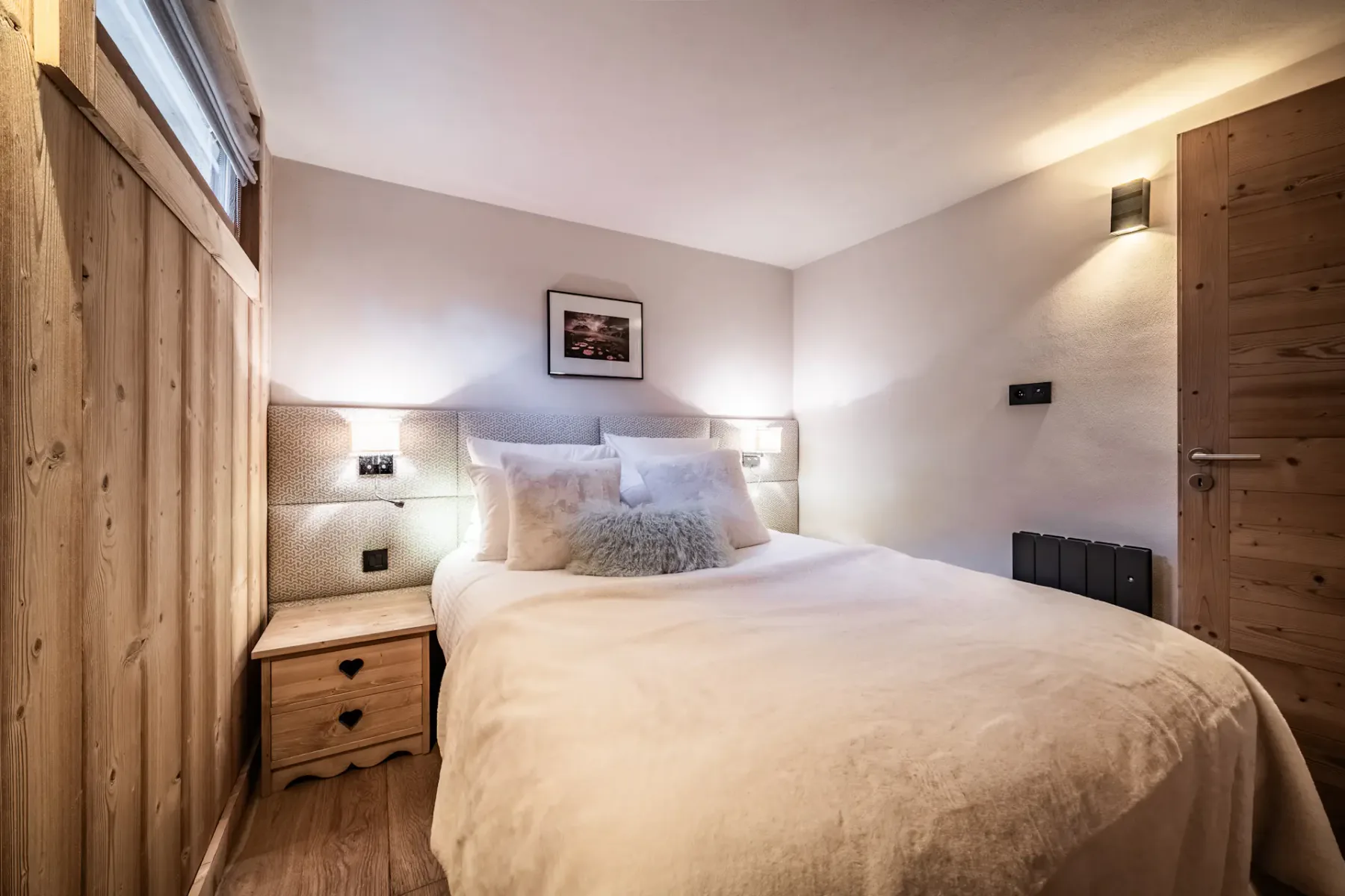 Cozy bedroom with a double bed, wooden walls, soft lighting, and a small window. A nightstand with a lamp is beside the bed.