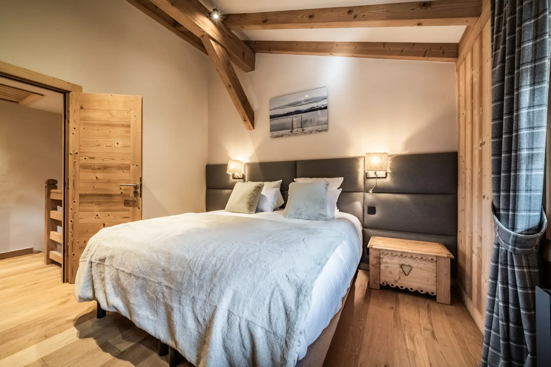 Cozy bedroom with wooden walls, a double bed with white bedding, a painting above the headboard, and a window with plaid curtains.
