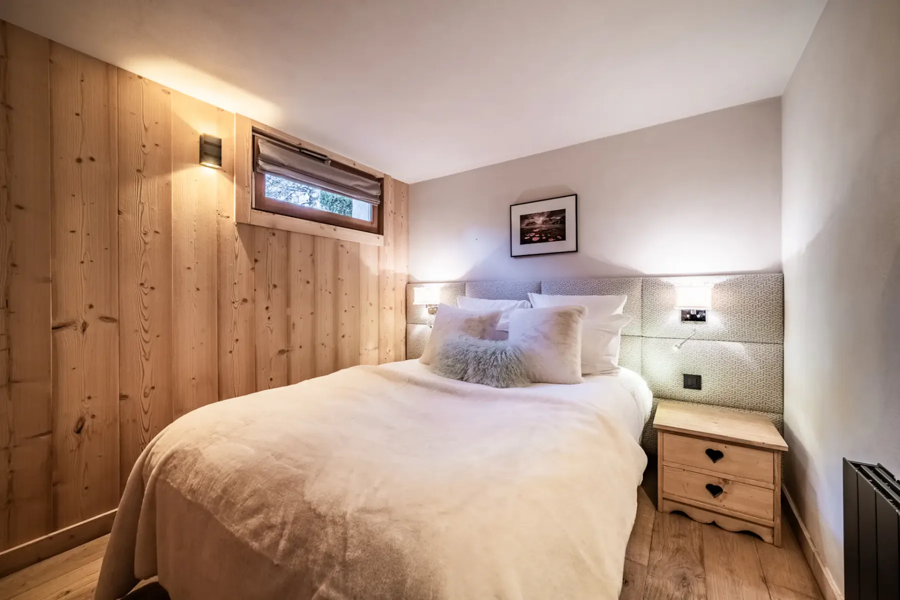 Cozy bedroom with a double bed, wooden walls, soft lighting, and a small window. A nightstand with a lamp is beside the bed.