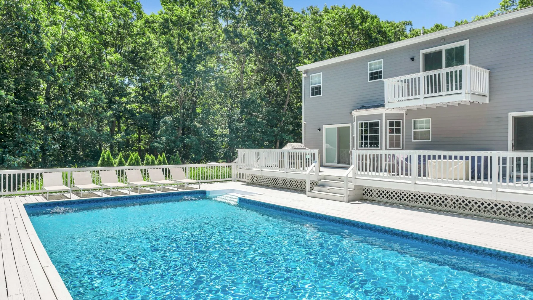 An outdoor swimming pool sits beside a gray house and white deck.