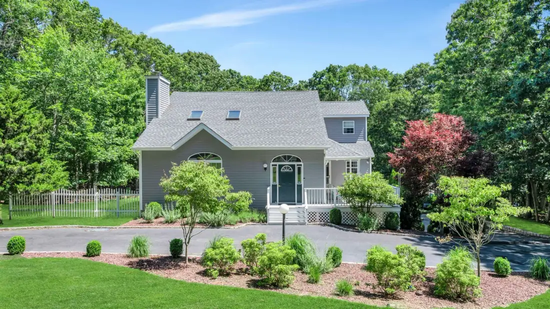 A gray house stands among green trees, with a lawn and landscaped garden in front.