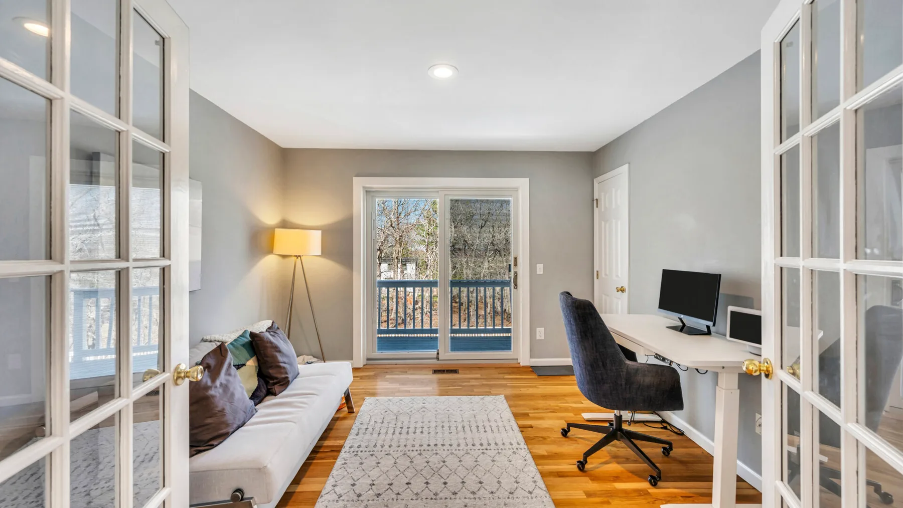 A bright home office with a desk, seating, and glass doors opening to a balcony.