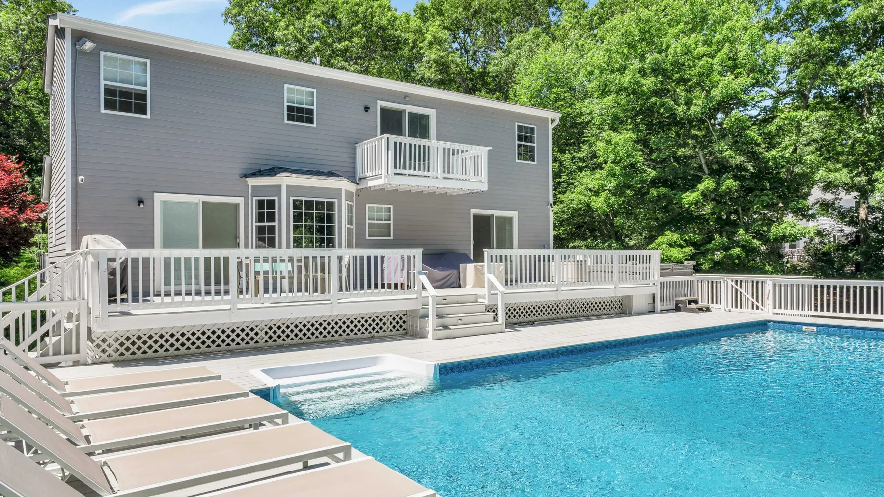 A swimming pool and raised deck sit beside a gray house surrounded by trees.