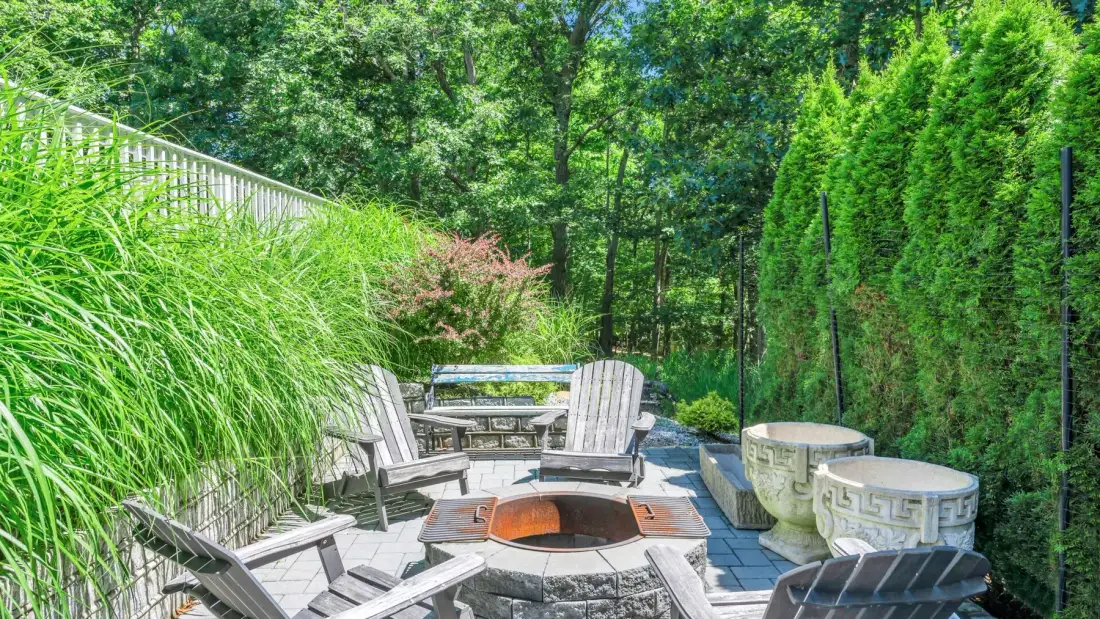 An outdoor patio has seating around a fire pit, with greenery along the edges.