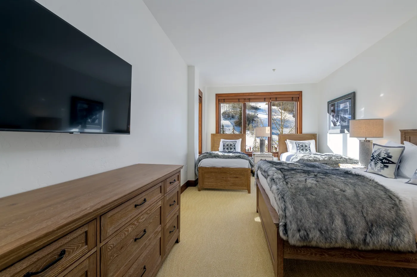 A bright bedroom with two beds, a wooden dresser, and a large window.