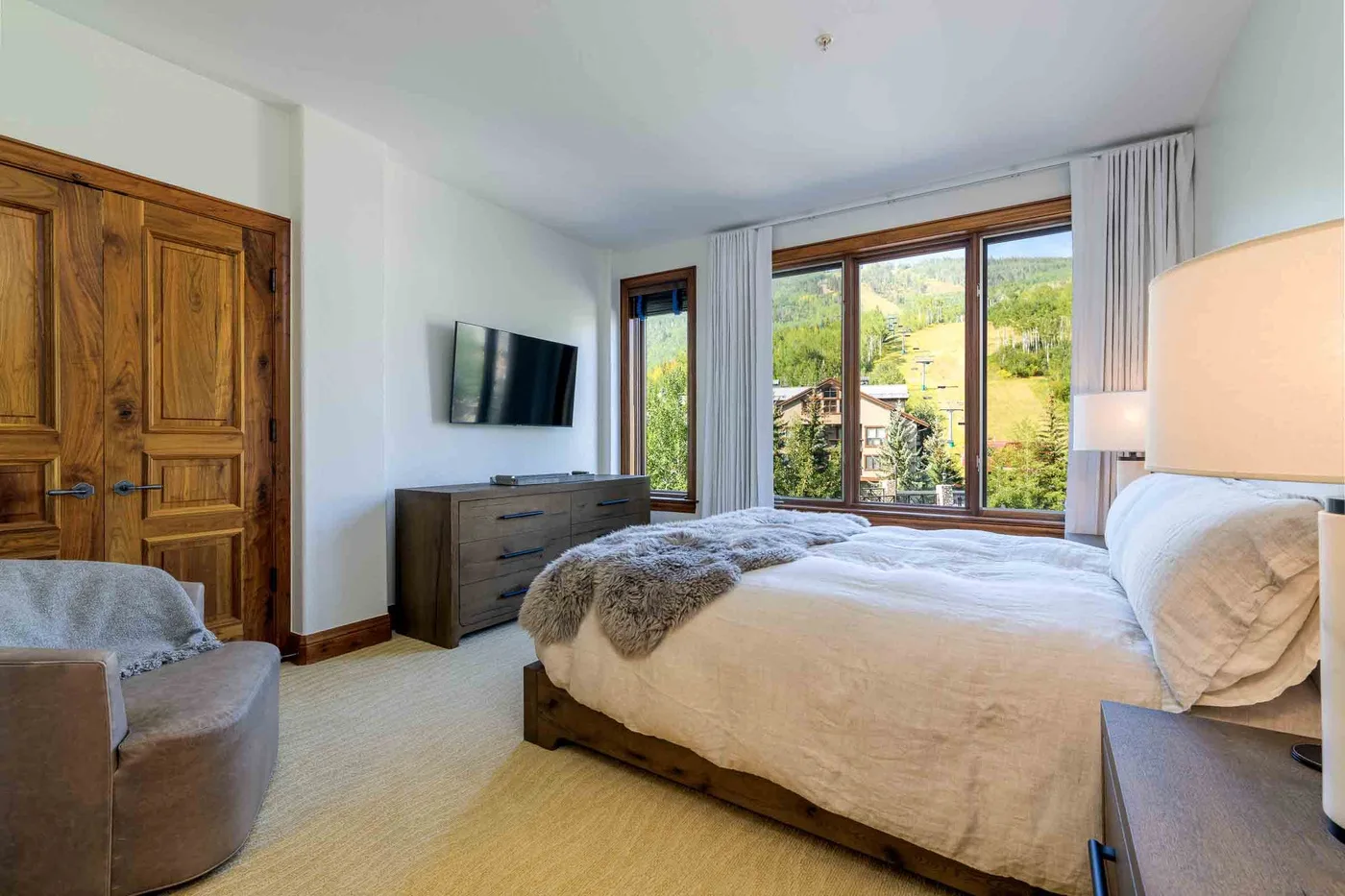 A bedroom with a large bed, wooden doors, and windows overlooking greenery.