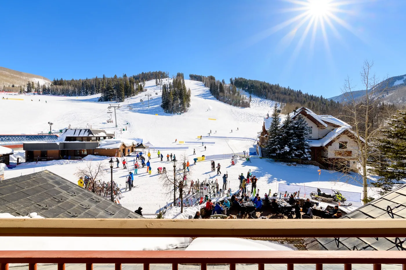 A sunny ski resort scene with snowy slopes and people gathered near the base.