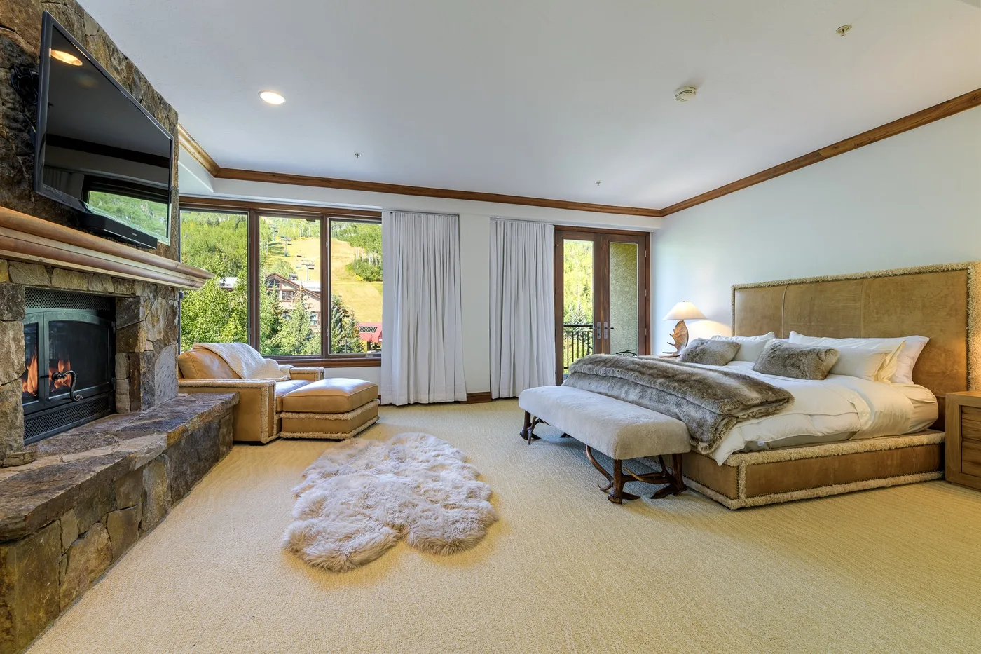 A bedroom with a large bed, a stone fireplace, and windows overlooking greenery.