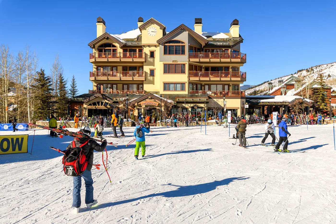 A large lodge stands beside a busy snowy ski area under a clear blue sky.