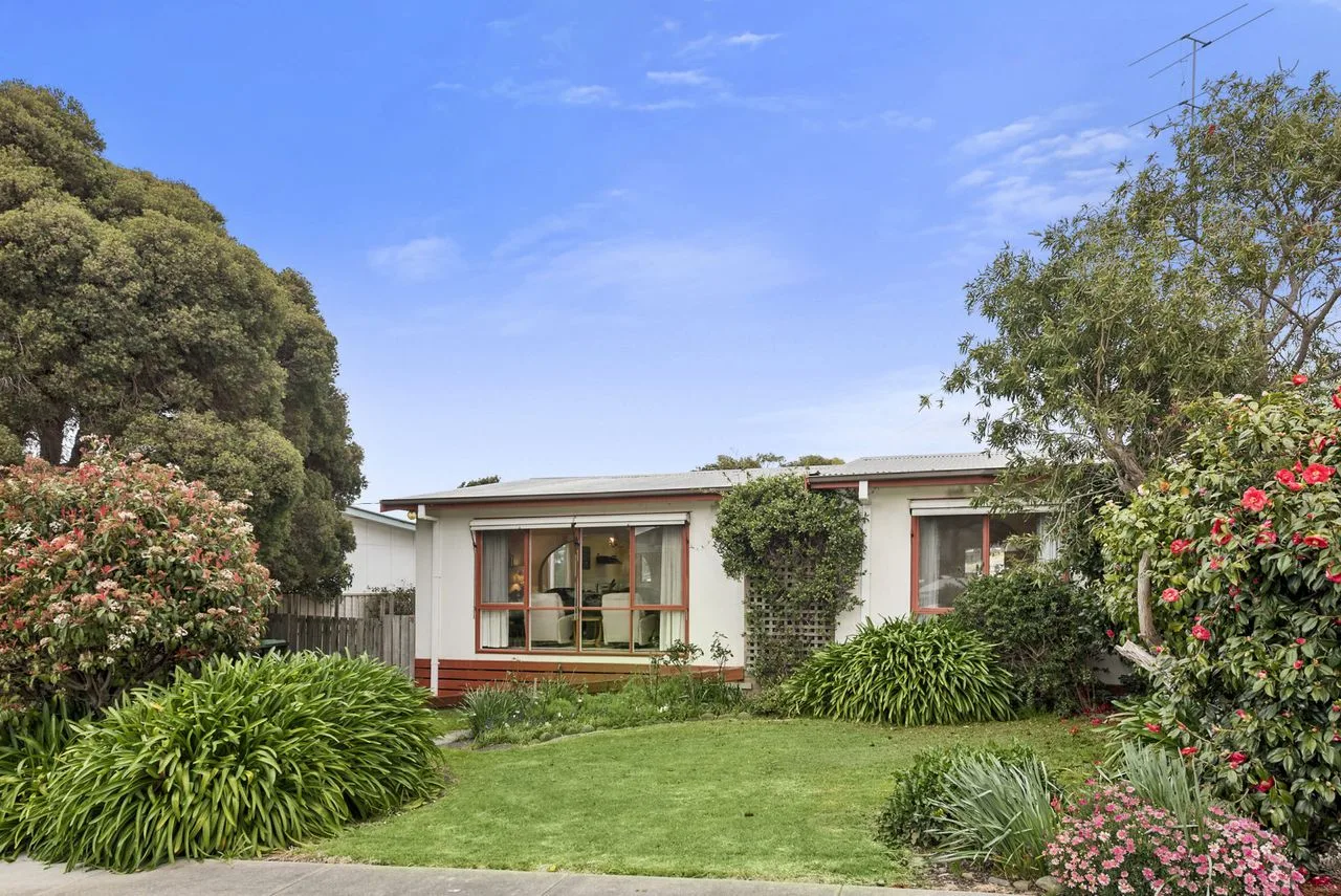 A single-story house stands behind a green lawn and flowering shrubs.