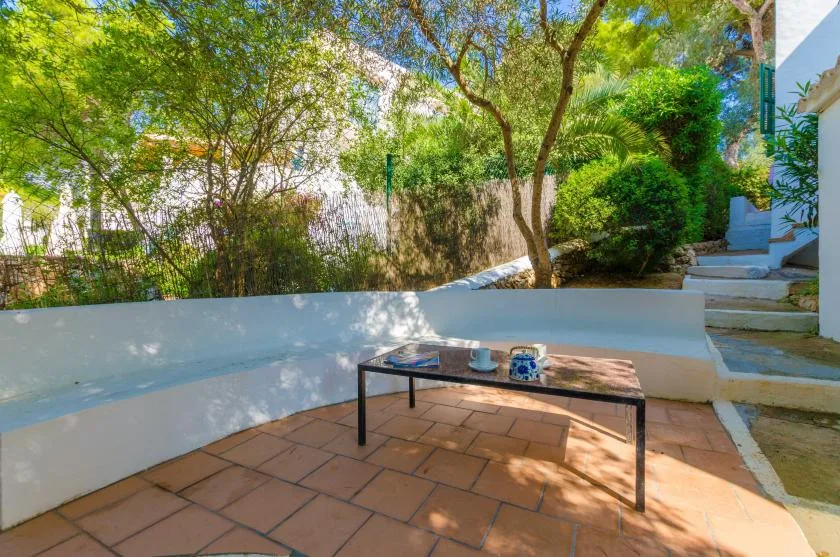Un patio exterior a la sombra con una mesa baja y vegetación alrededor.
