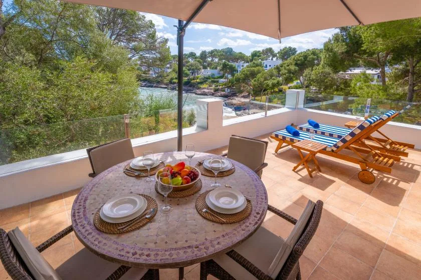 Una terraza exterior con comedor da a la costa, con una mesa a la sombra y tumbonas.