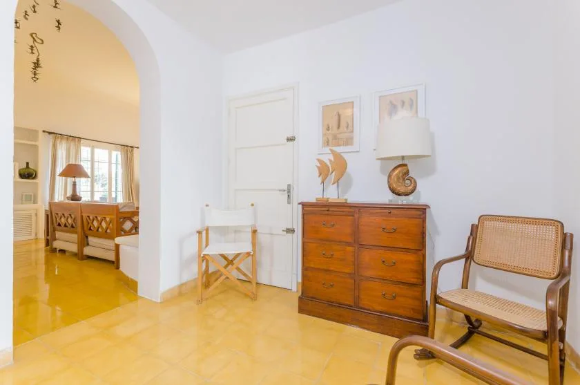 Una habitación luminosa con una cómoda de madera, cuadros y un arco que da a una sala de estar.