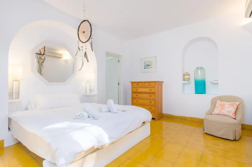 Un dormitorio luminoso con una cama bien arreglada y una silla acolchada.