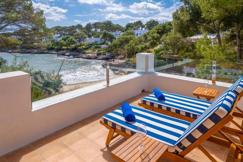 A sunny terrace overlooks the sea, with striped loungers and trees along the shore.