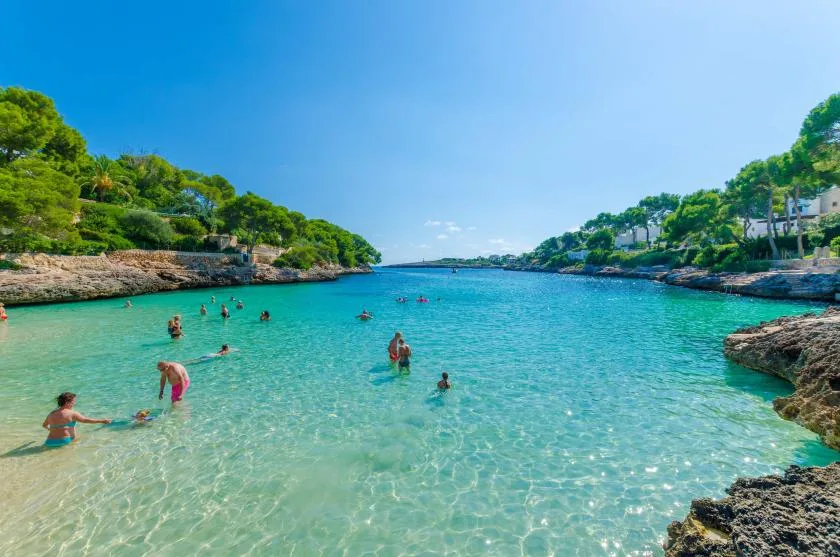 Una cala de aguas turquesas y claras se extiende entre orillas rocosas y árboles verdes.