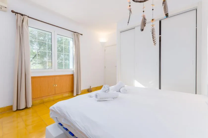Un dormitorio luminoso con ropa de cama blanca, ventanas con cortinas y suelo amarillo.