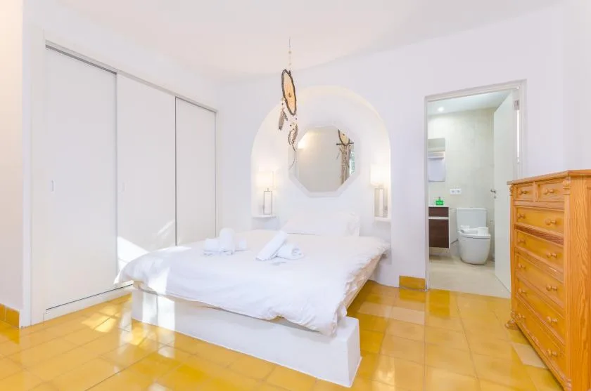 Un dormitorio luminoso con cama blanca, nicho de pared con espejo y puerta abierta al baño.