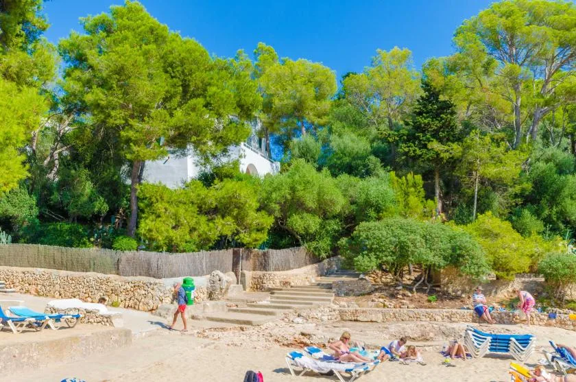 Una playa de arena con tumbonas se encuentra bajo terrazas de piedra y una ladera arbolada.