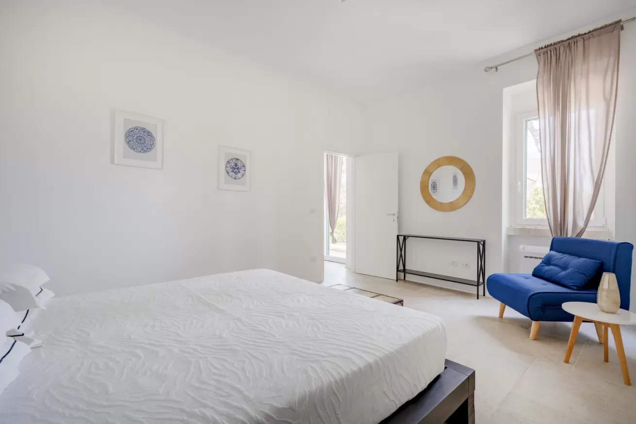 A bright bedroom with a large bed, blue seating, and tall windows.