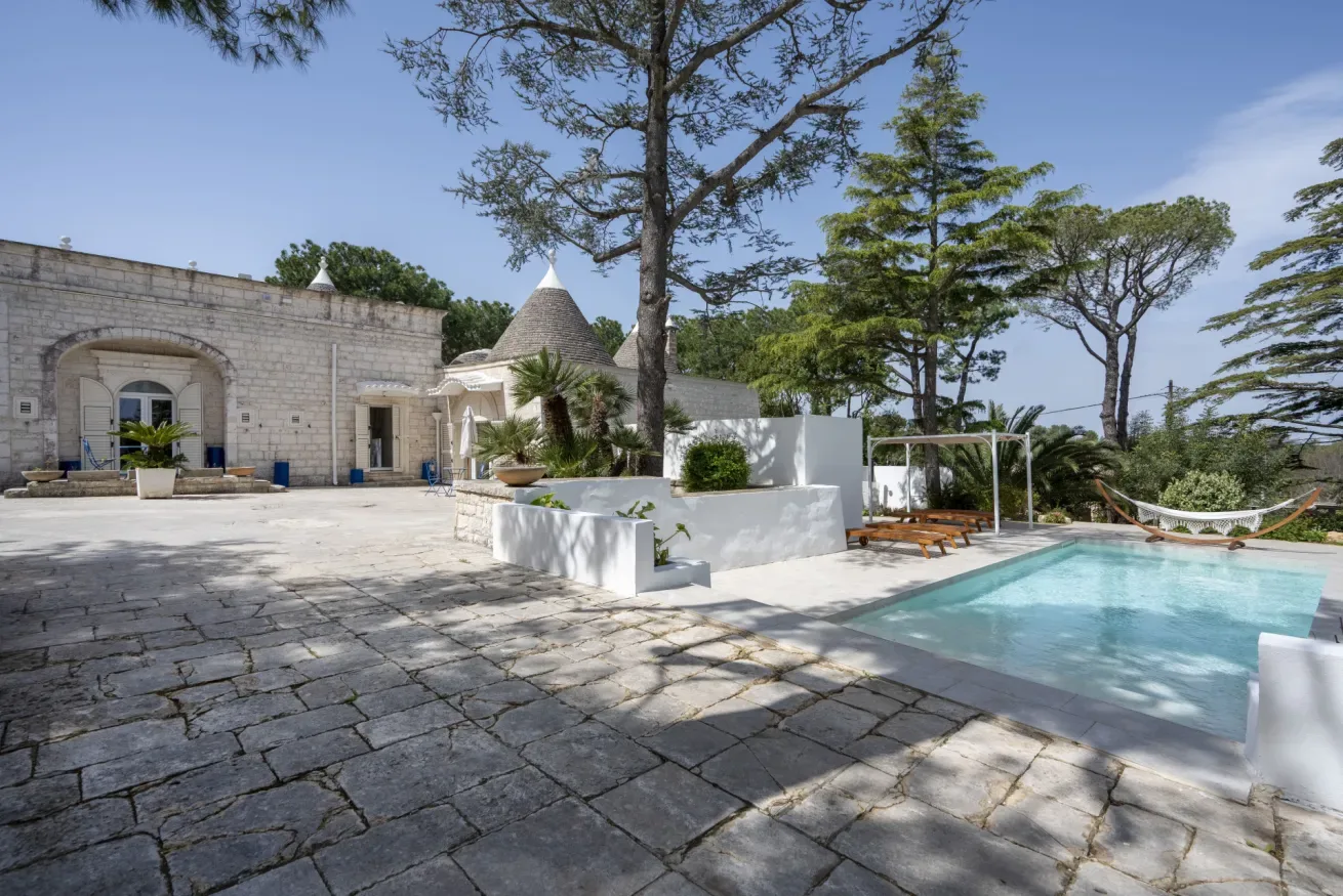A stone courtyard beside a swimming pool, with trees and outdoor seating.