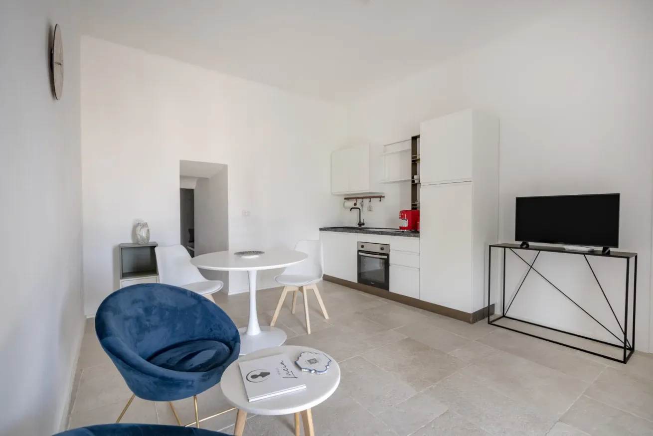 A bright open-plan room has a compact kitchen, dining table, and television.