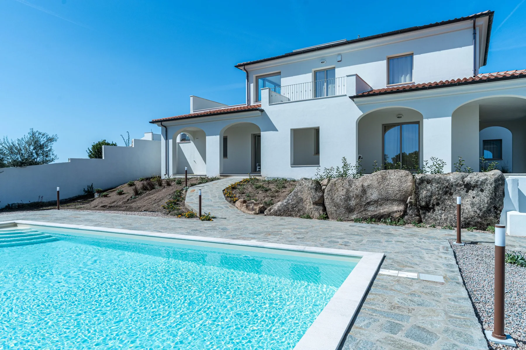 A modern white house overlooks a swimming pool and paved terrace.