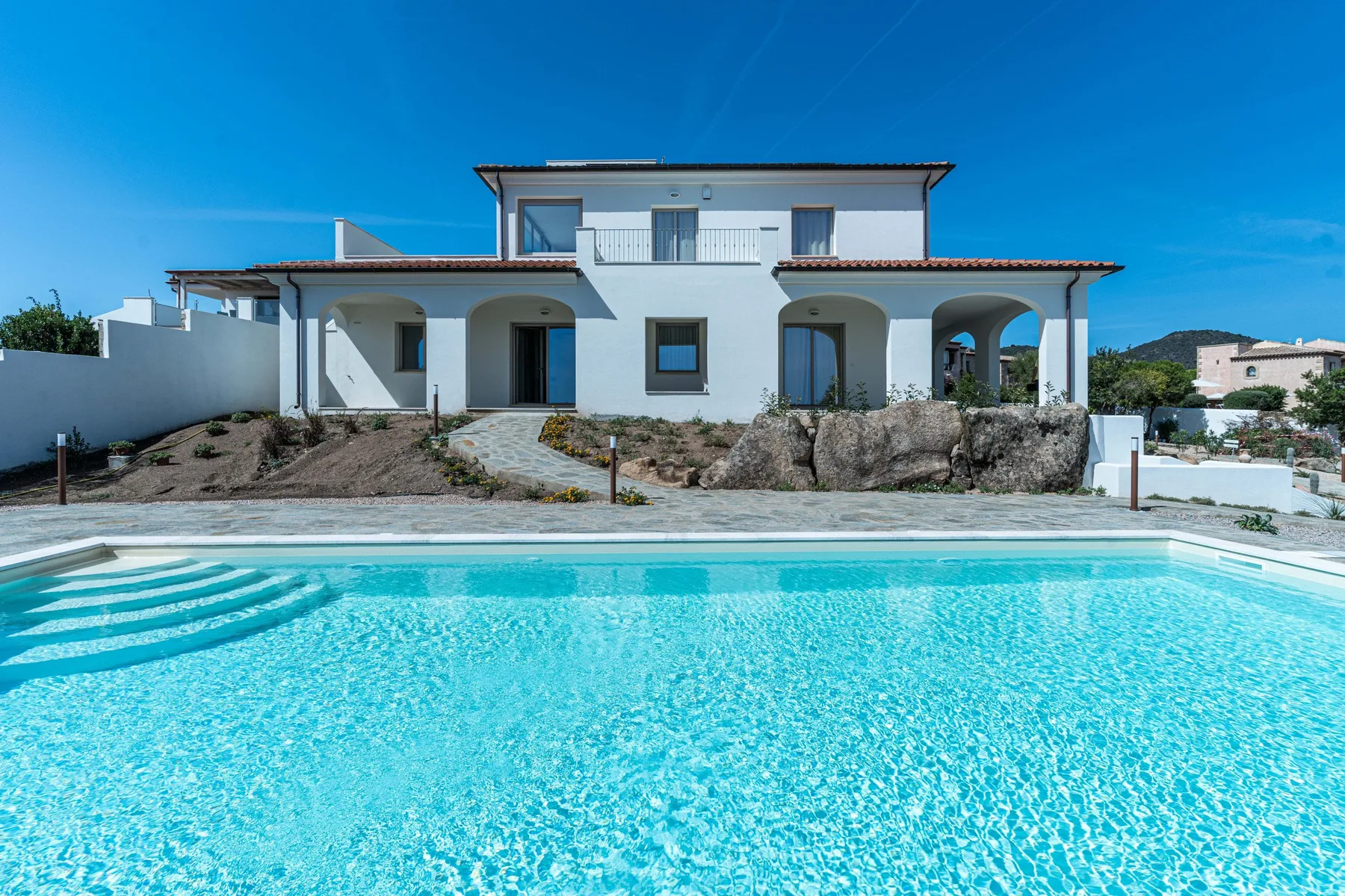 A white two-story house stands behind a bright blue swimming pool.