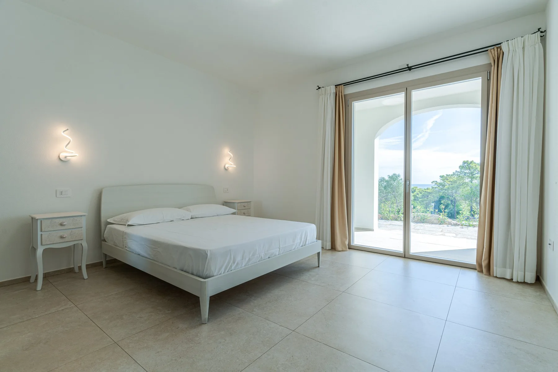 A bright bedroom with a bed beside a large glass door opening onto greenery.