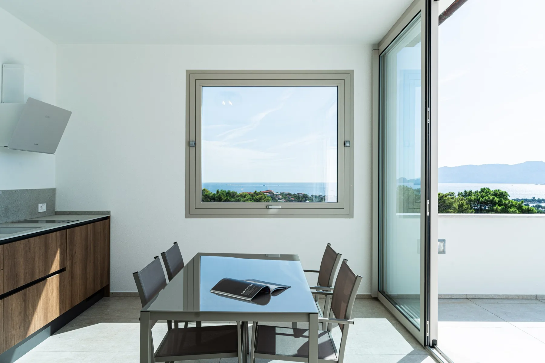 A bright dining area with a glass table beside a large window overlooking the water.