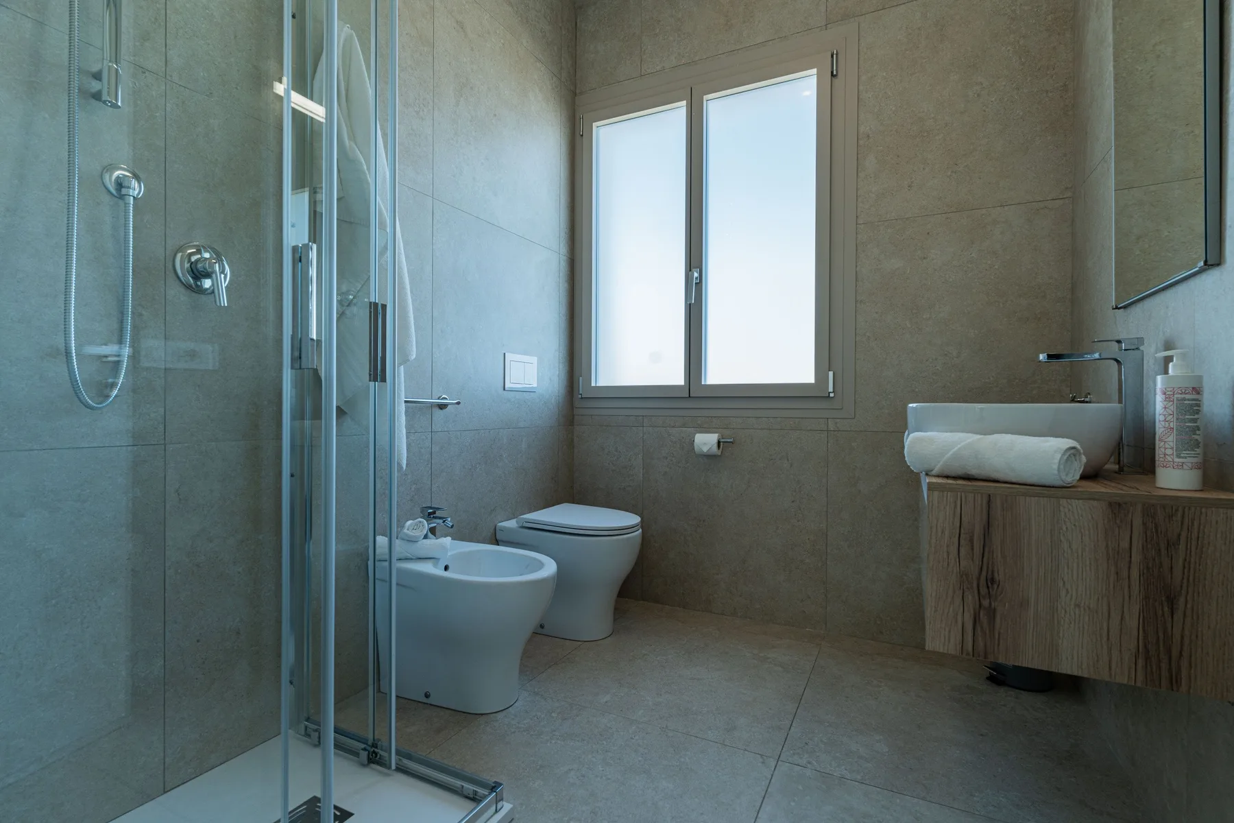 A tiled bathroom with a glass shower, toilet, and sink beneath a window.