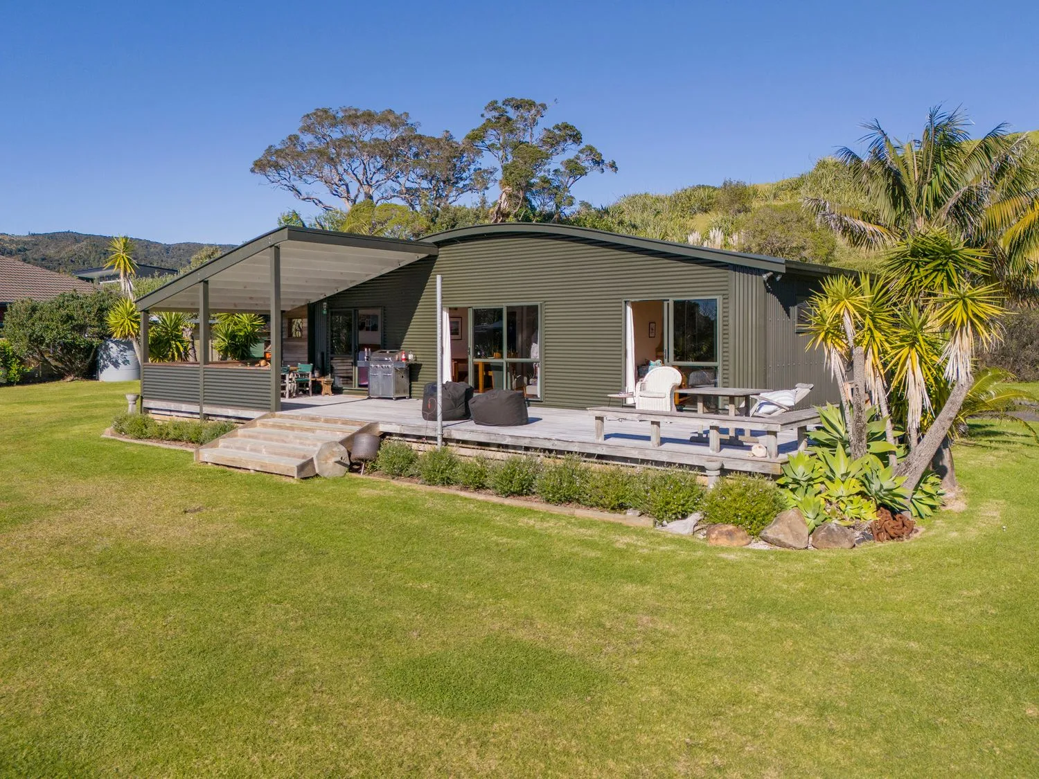 A single-story house with a covered porch and outdoor seating on a wooden deck surrounded by a lawn and trees at Wai Whare - Kuaotunu Holiday Home in Kuaotunu