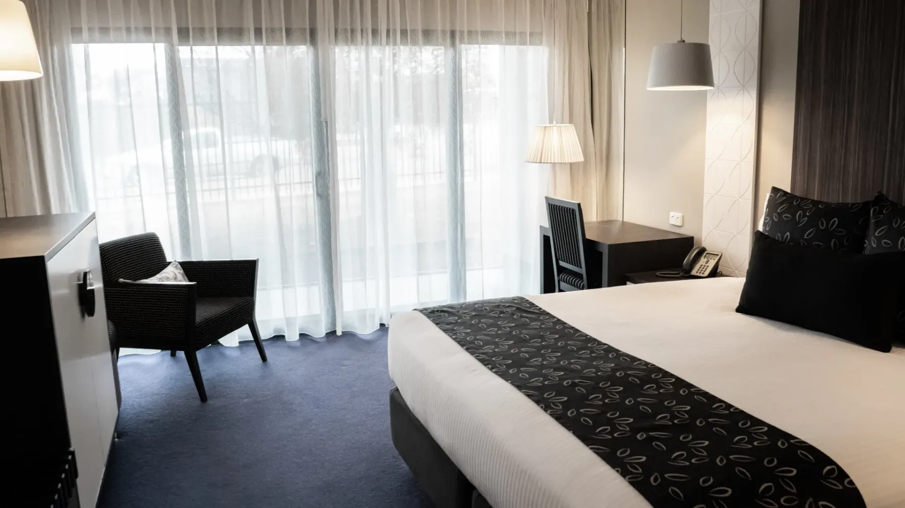 Habitación de hotel con una cama, silla, escritorio y puerta de cristal.