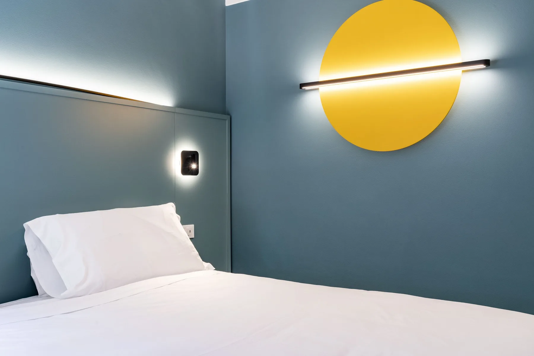 A bedroom with white bedding and a round yellow wall light.