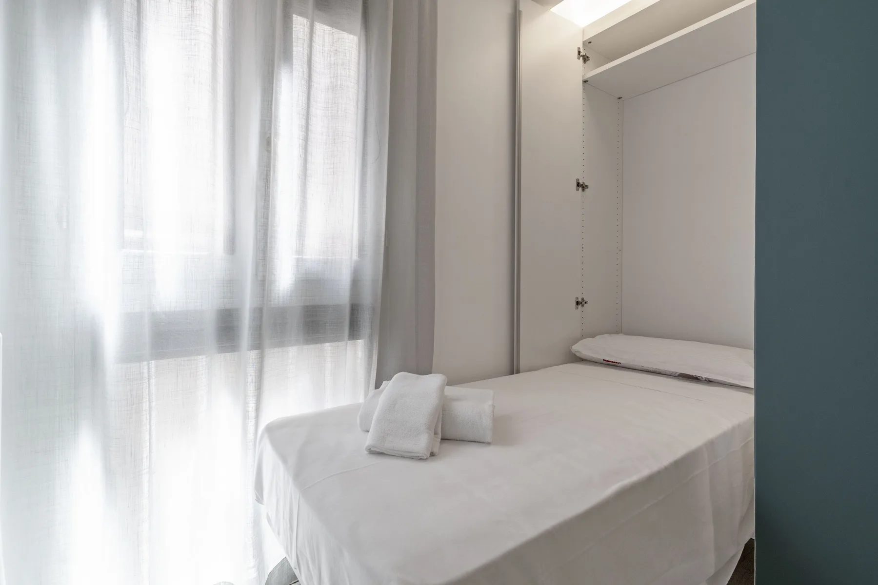 A bedroom with a bed beside a bright window with sheer curtains.
