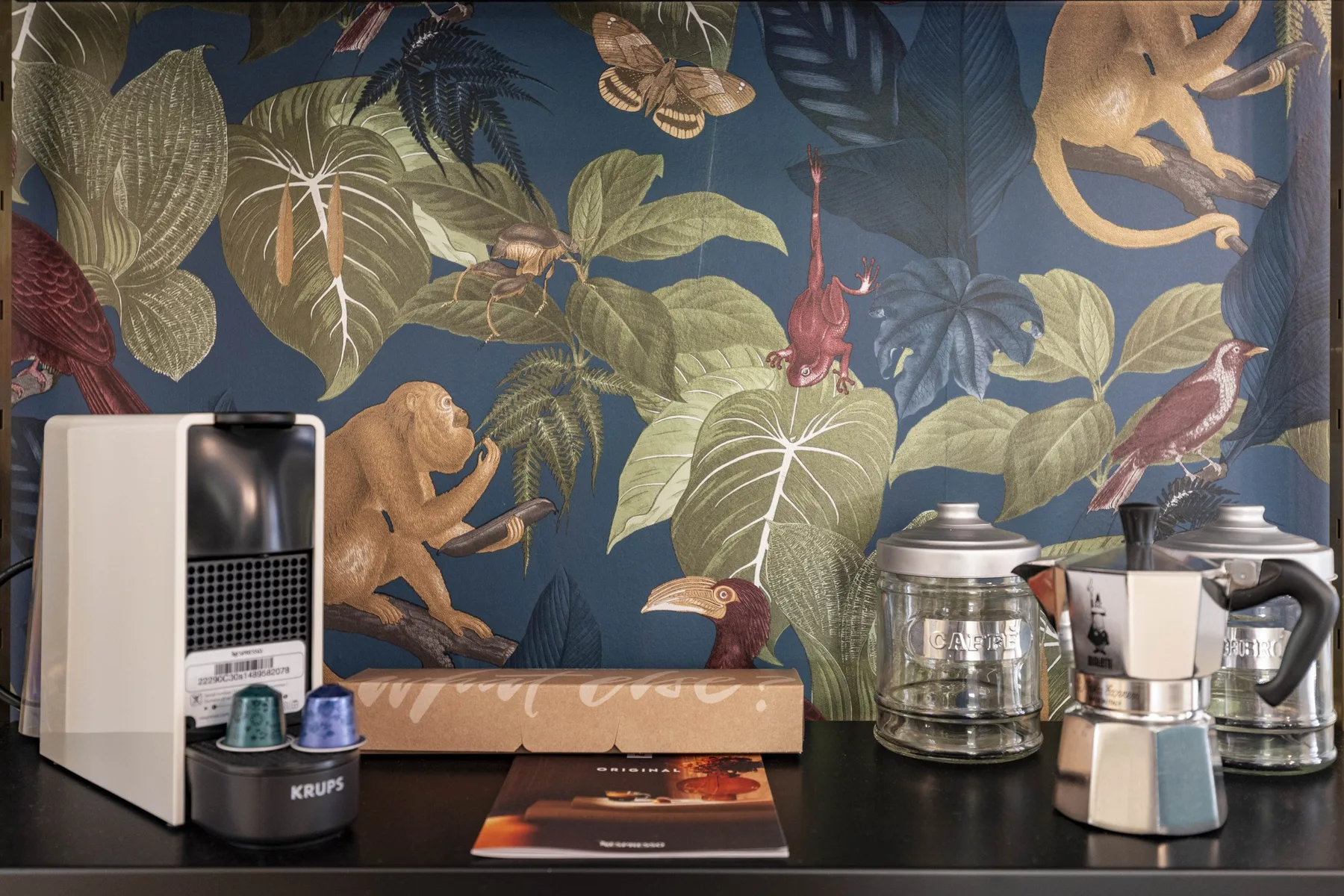 A kitchen counter with coffee-making appliances sits against leafy animal-patterned wallpaper.