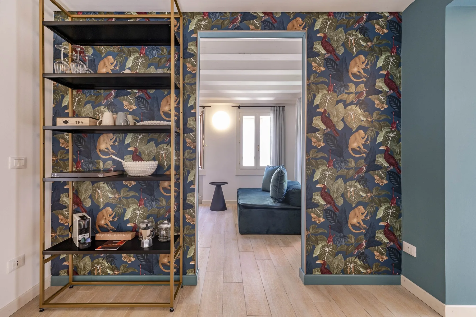 A wallpapered living area with open shelving and a view into a room with seating.