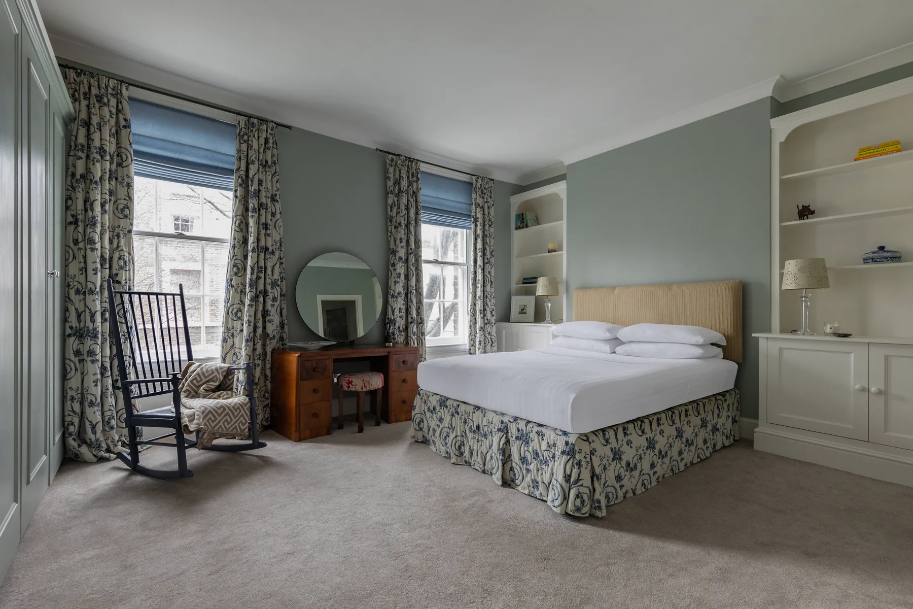 A bedroom with a made bed, a rocking chair, and tall windows.