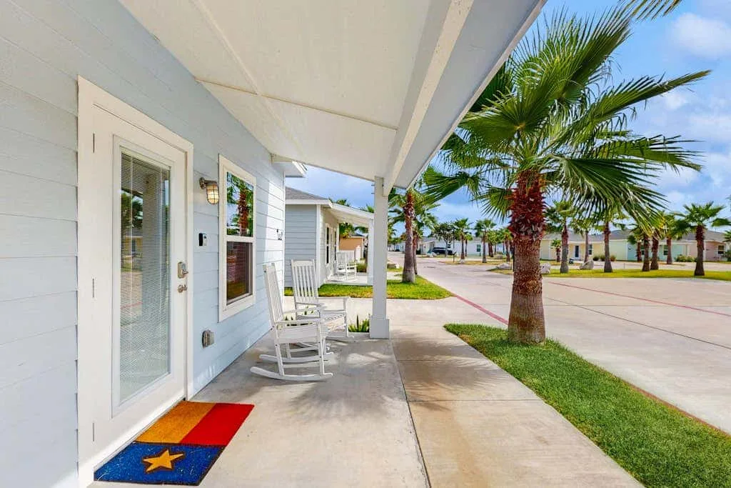 A covered porch with rocking chairs overlooks a sidewalk and palm trees.