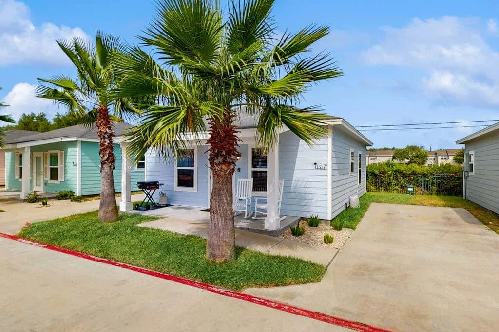 A small white cottage sits beside a paved driveway, with palm trees in front.